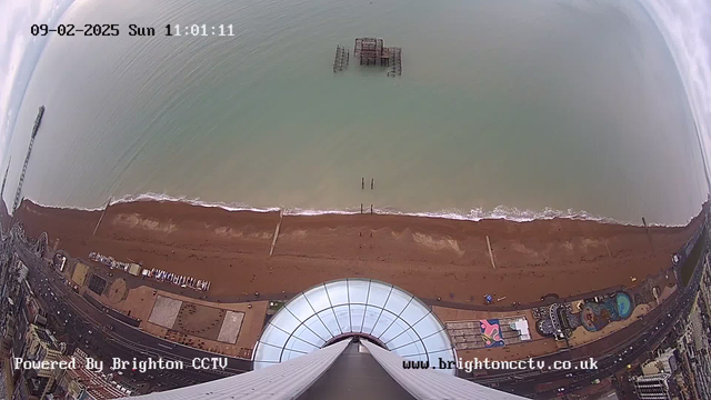 A high-angle view from a webcam overlooking a sandy beach and the sea. The shoreline is visible with gentle waves lapping at the sand. In the water, there is an old, partially submerged pier structure. The beach area appears mostly empty, with a few people and colorful beach umbrellas scattered near the water. A portion of the boardwalk and some attractions can be seen in the lower part of the image, alongside parts of the city. The sky is mostly cloudy, and the image has a timestamp indicating the date and time.