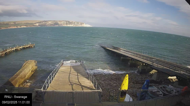 A view of a coastal area featuring a calm sea with gentle waves. In the foreground, there's a wooden ramp leading down to the water, surrounded by a metal railing. To the left, a solid structure extends into the water. Several colorful kayaks, including yellow and blue ones, are positioned on the shore. In the background, cliffs and rolling hills are visible under a partly cloudy sky.