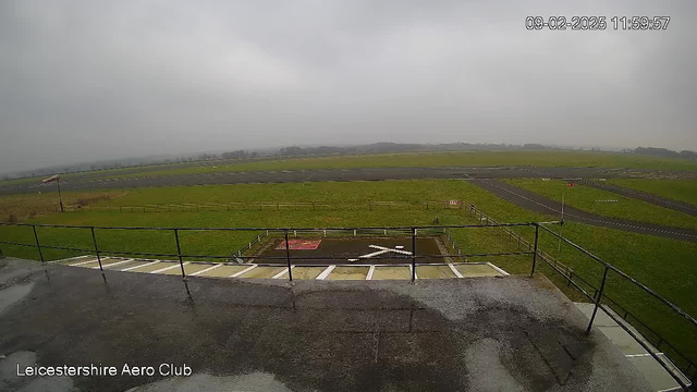 A view from a webcam positioned at the Leicestershire Aero Club, showing a large grassy area and an airfield under overcast skies. The ground is wet, likely from recent rain, and there is a low railing in the foreground. A runway is visible, with markings and several signs along its edges. In the distance, there are trees and a hint of a horizon, but the scene has a muted light due to the cloudy weather.