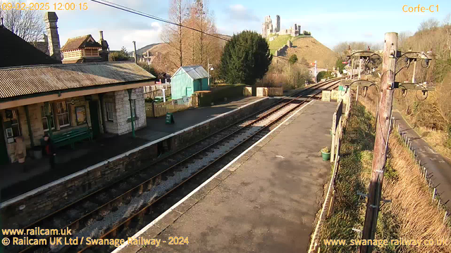 A railway station platform with two sets of railway tracks running parallel. In the foreground, there is a stone platform with a green bench and a sign that reads "STAIR OUT." On the left, a brick building with a sloped roof and a chimney. To the right, there is a blue shed with large windows. In the background, a hill features a historic castle with several towers. The sky is mostly clear with some clouds. The scene is illuminated by natural sunlight.