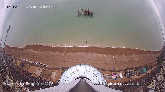 A bird's-eye view of a sandy beach meeting the ocean, with gentle waves lapping at the shore. The shoreline has a mix of wet sand and dry areas, and a few people can be seen walking near the water. In the distance, there is a partially submerged structure, likely a pier or ruins, on the horizon. The foreground features a large circular structure, possibly a viewing platform or attraction. The scene is captured in the daylight under a clear sky.