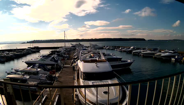 A view of a marina filled with boats docked along wooden piers. The scene is bathed in sunlight, with a partly cloudy sky overhead. Several yachts and smaller vessels are moored in the calm water, and a distant green landscape is visible across the bay. The image captures a tranquil and serene coastal atmosphere.