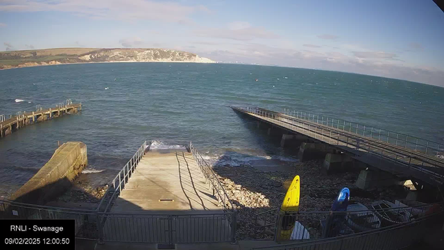 A coastal scene featuring a calm sea and a clear blue sky. In the foreground, there is a docking area with steps leading down to the water. To the right, two kayaks—one yellow and one blue—are positioned near the shore. A wooden pier extends into the water on the left side, with a rocky beach visible at the bottom. In the background, a line of cliffs can be seen, suggesting a scenic landscape. The image captures a peaceful outdoor environment.