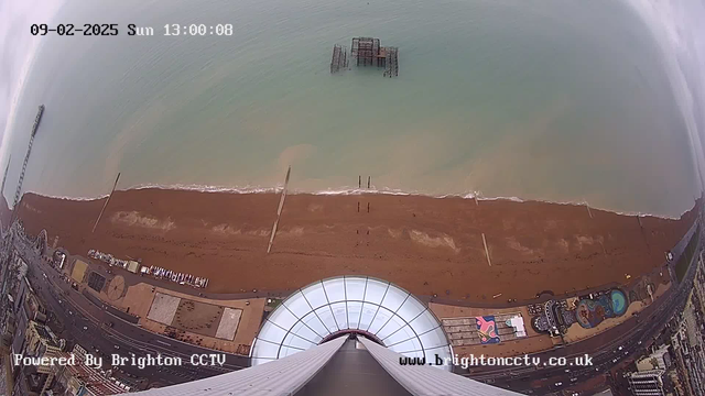 Aerial view of a beach with brown sand and calm sea, captured from a high point. The image displays a partially submerged pier structure in the water. On the beach, there are scattered people and objects, and a promenade lined with buildings and attractions is visible in the lower section of the image. The sky is overcast, creating a muted atmosphere. The time and date are displayed in the top corner, showing Sunday, February 9, 2025 at 1:00 PM.