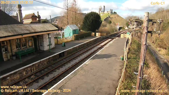 A railway station platform with tracks running through it. To the left, there is a stone building with a sloping roof, possibly a waiting area, and a green bench in front. A small, teal-colored shed is visible beside a wooden fence, which has a sign that reads "Way Out." In the background, there is a hillside crowned by a castle ruin. The sky is partly cloudy, with some blue peeking through. The scene is bright and well-lit.