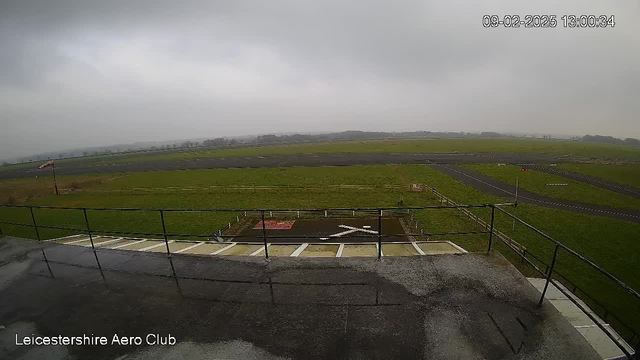 A cloudy sky looms over a green field and a runway at Leicestershire Aero Club. The foreground features a balcony railing, with wet pavement visible. In the center of the image, there is a marked area on the ground with a red mat and a white cross. In the distance, the runway stretches out, bordered by a wooden fence. No aircraft are visible in the scene.