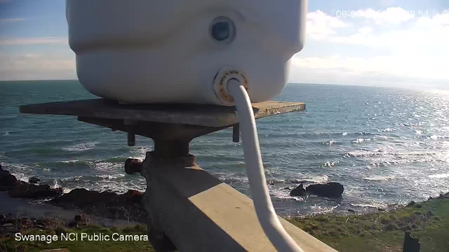 A white container is positioned on a metal platform overlooking the sea. The container has a visible pipe extending from it. Below, the waves of the ocean crash against rocky shorelines. The scene is bathed in bright sunlight, reflecting off the water, with a clear sky and scattered clouds in the background.