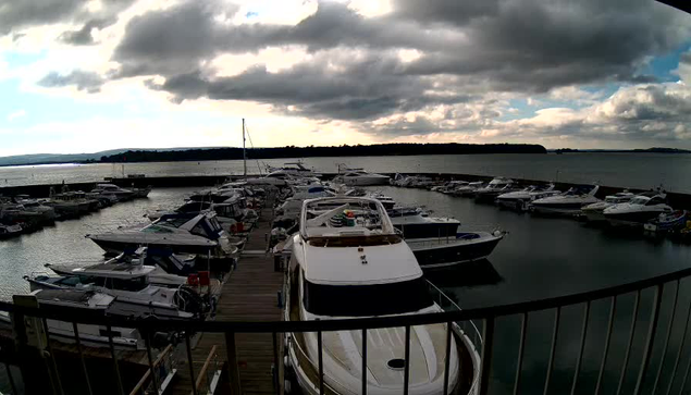 A marina filled with various boats docked along wooden piers. The scene is set under a cloudy sky with some hints of sunlight peeking through. The water reflects the boats and the clouds above, creating a tranquil atmosphere. In the background, distant land can be seen along the horizon.