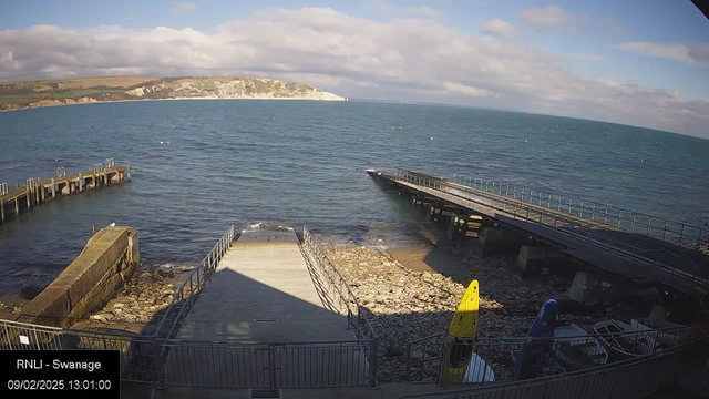 A seaside view showing a calm blue ocean under a partly cloudy sky. In the foreground, there are two wooden piers; one extends into the water, and the other leads down near a rocky shoreline. On the shore, there are scattered stones and some visible equipment, including a yellow kayak. The scene is bright and inviting, depicting a tranquil coastal environment.