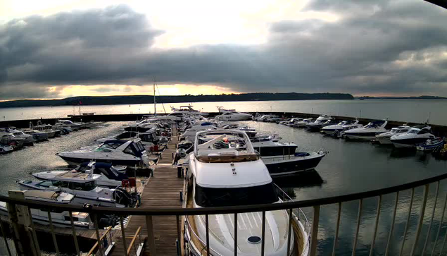 A view of a marina filled with numerous boats docked along a wooden walkway. The water is calm and reflects the cloudy sky, which has patches of sunlight breaking through. In the background, hills can be seen along the horizon, while the scene is framed by a railing in the foreground.