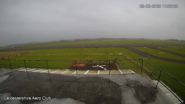 A cloudy, overcast sky is visible in the background. In the foreground, there is a grassy field with a small section of pavement. A windsock on a pole indicates light wind direction. An area marked with a white cross is visible on the ground, alongside a red rectangle. There are white railings at the edge of the viewing platform, which is wet, suggesting recent rain. The image is labeled with the name "Leicestershire Aero Club" in the bottom left corner, along with a timestamp.