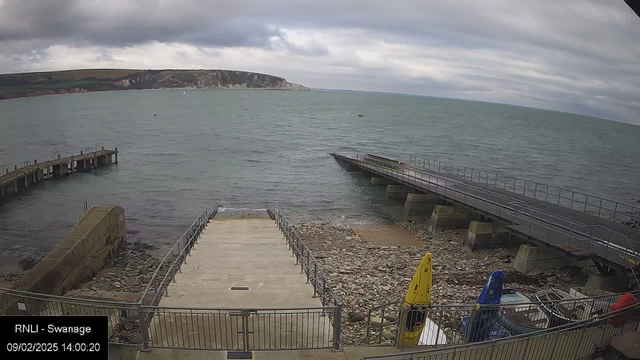 A view of a seaside location featuring a light gray sky with scattered clouds. The water is a calm blue-green, and there are distant cliffs visible on the horizon. In the foreground, a set of cement steps leads down to the water, bordered by a metal railing. To the left, a wooden pier extends into the water, and on the right, a wide ramp leads down to the water's edge. There are kayaks in yellow, blue, and red resting against a stone wall. The area is rocky, with some visible stones on the ground near the water.