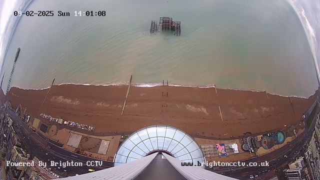 Aerial view of a beach. The sandy shore is visible with gentle waves lapping at the edge. Several wooden pier structures extend into the water, and there are remnants of an old pier partially submerged. Along the beach, a row of paths and structures line the shore. The image is taken from a high vantage point, looking down towards the beach and ocean, with clouds in the sky above. An observation tower base is seen at the bottom of the image.