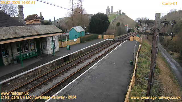 A railway station scene featuring a stone building with a green roof and a covered area on the left. A long platform with two sets of railway tracks runs through the middle. The background shows a hill with castle ruins on top, surrounded by trees. A sign on the platform indicates "WAY OUT," and there are wooden fences along the edges, with a small green building to the left. The sky is overcast.