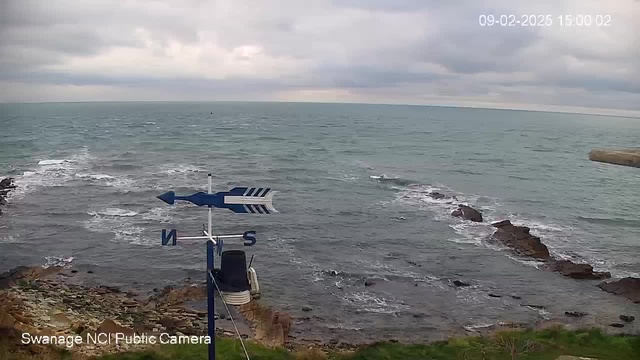 A coastal scene with a cloudy sky and a gray ocean. The water has small waves and some visible rocks in the foreground. In the lower left, there is a weather vane displaying the directions 'N' (north) and 'S' (south). The shore is rocky, and some grass is visible near the edge of the water. The image is timestamped in the upper right corner.