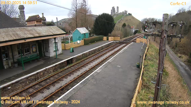 A vacant railway station platform with a stone building on the left featuring a peaked roof and a green bench. In the background, there is a turquoise shed and a wooden fence with a sign that reads "WAY OUT." The railway tracks run parallel to the platform, and in the distance, a hill can be seen with remnants of an old stone castle. The sky is cloudy and overcast. A wooden utility pole with wires stands on the right side of the image.