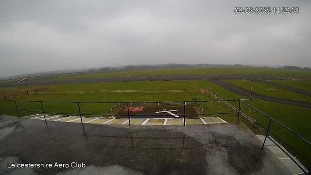 An outdoor view from a raised platform at an Aero Club, looking over a grassy runway and landing area. The sky is overcast and gray, casting a dim light over the scene. A wooden fence lines part of the runway, and a single windsock is visible on the left side. In the foreground, there is a white X marked on the ground, possibly indicating a landing zone. The runway appears to be clear of any aircraft or activity.