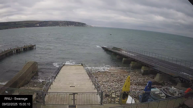 A view of the sea from a webcam, featuring a rocky shoreline and cloudy skies. On the left, there is a wooden pier extending into the water. In the foreground, a concrete ramp leads down to the water, flanked by metal railings. To the right, a second pier juts out, with some kayaks in yellow, blue, and orange resting on the shore. The water appears calm, with gentle waves lapping at the rocks.