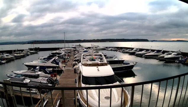 A marina scene featuring numerous boats docked along a wooden pier. Various sizes of boats are visible, some covered with blue and white tarps. The water is calm, and the sky is overcast with gray clouds. In the background, there is a hint of land with trees, and the horizon line is visible where the water meets the sky.