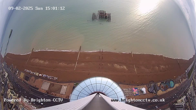 Aerial view of a beach and coastline during the day. The foreground shows a circular structure with a glass dome, leading down to a sandy beach with scattered people and some wooden posts in the water. An old, partially submerged pier can be seen further out in the sea. To the left, part of an amusement park and a road lined with buildings are visible. The ocean has a calm look, reflecting the sunlight, while the sky is mostly clear. The timestamp at the top indicates it is Sunday, February 9, 2025, at 15:01:12.