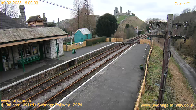 A view of Corfe Castle railway station, featuring a stone building with a steeply pitched roof and several windows. In the foreground, there are two sets of railway tracks running parallel. To the left, a long green bench is situated beside the platform, and a sign indicates "Way Out." Behind the station, a small blue shed is visible. In the background, Corfe Castle ruins sit atop a hill, surrounded by trees and a cloudy sky. The scene is set in a rural location, with a path and a footpath leading away from the station.