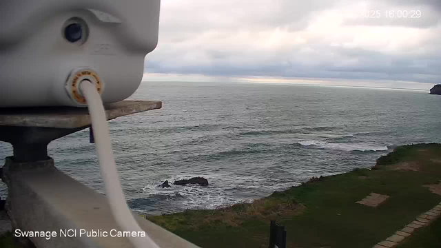 A view of the ocean under a cloudy sky, with waves gently lapping against rocky formations. In the foreground, there is a white object attached to a metal structure, possibly part of a camera or monitoring equipment. The shoreline is visible to the right, with grass and stone pathways leading down to the water. The overall atmosphere is calm and tranquil.