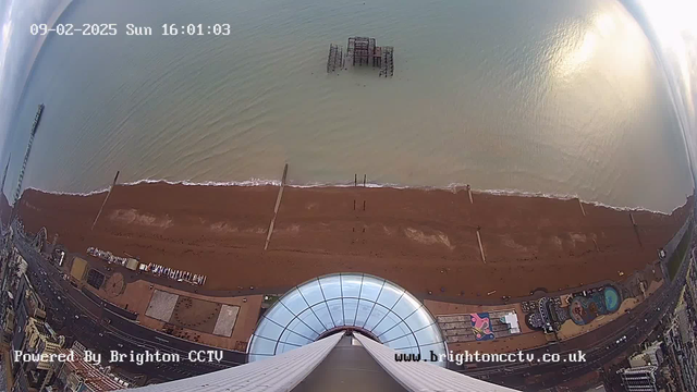 A high-angle view of a beach and coastline, with sandy shores extending towards the ocean. The water is calm, glistening under sunlight, and several wooden piers extend into the sea. In the distance, a dilapidated structure is partially submerged in the water. Below, there are amusement rides and vibrant murals on the ground near the edge of the beach. The scene captures a mix of natural beauty and human development, with data displayed indicating the date and time.