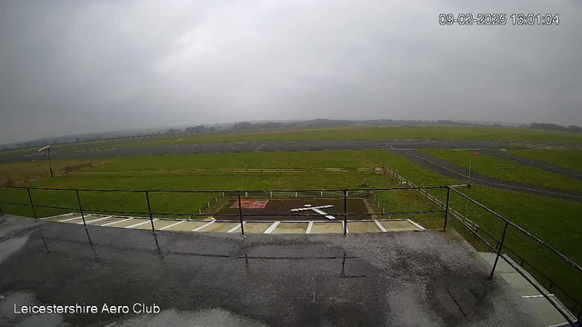 A cloudy scene from a webcam located at Leicestershire Aero Club, showing a grassy area with a runway in the background. In the foreground, there is a flat roof with railings, and a white cross marked on the ground. The sky appears gray and overcast, with gentle winds indicated by a nearby windsock. The area is mostly empty, with a distant view of trees on the horizon.