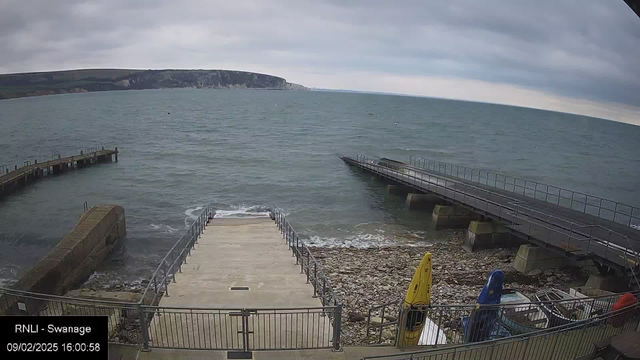 A view of the coastline with a rocky shore leading to the sea. In the foreground, a concrete ramp descends to the water, flanked by railings. To the left, there is a wooden pier extending into the waves, while another pier extends from the right side of the image. Several kayaks are stored on the shore, with colors including yellow, blue, and red. The sky is overcast with clouds, and the water appears choppy. In the background, cliffs can be seen along the horizon. The date and time are displayed in the lower left corner.