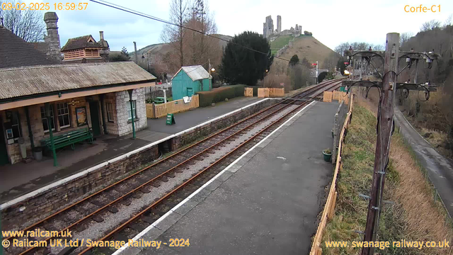 A view of Corfe Castle railway station. The foreground features a stone platform with a green bench on the left and a sign reading "Way Out." In the background, a single railway track runs towards the right. The station building has a sloped roof and several windows. To the right, a weathered wooden utility pole with wires stands beside the track. On the hill behind the station, the ruins of Corfe Castle are visible, surrounded by trees and grassy slopes under a cloudy sky.