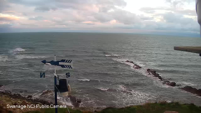 A scenic view of the ocean taken from a webcam. In the foreground, a blue and white weather vane indicates direction, with the letters "N" and "S" visible. The ocean stretches out toward the horizon, with gentle waves and some rocky formations visible along the shore. The sky is partly cloudy with soft pastel colors from the setting sun. The overall atmosphere feels calm and serene.