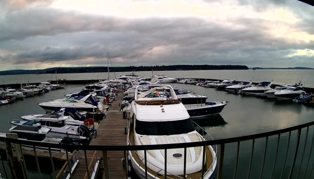 A marina filled with various boats docked at a wooden pier. The scene includes multiple white and blue boats, some larger yachts, alongside a calm body of water. In the background, there are dark, cloud-covered skies and a distant shoreline with greenery. The atmosphere appears tranquil, with soft light reflecting off the water.