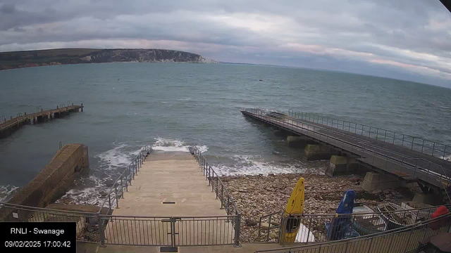 A view of a coastal scene featuring a calm sea with gentle waves. In the foreground, there is a set of stairs descending to the water, surrounded by railings. To the left, a rocky area with scattered stones is visible. There are two piers extending into the water, one made of wood and the other of concrete. On the right side, several colorful kayaks are stacked or resting near the shore. The sky is overcast with shades of gray, suggesting an impending change in weather, and a distant cliff can be seen along the shoreline in the background.