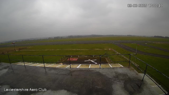 A cloudy sky looms over a wide-open field at an airport. The foreground shows a railing along the edge of a viewing platform. Below, a large white cross is marked on the ground, surrounded by a grassy area. In the distance, the airstrip extends, bordered by patches of greenery, with several lanes and a small windsock visible to the left. The atmosphere appears overcast and dull, indicating possible rain. The image timestamp is 17:00:49 on February 9, 2025.