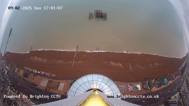 An aerial view of a beach at Brighton. The foreground features a circular structure, likely a viewing platform, with a glass top and visible support cables. Below, the sandy beach extends horizontally with gentle waves reaching the shore, and some wooden posts sporadically dotting the waterline. In the distance, an old pier structure is partially submerged in the water. The scene is set under a clear sky, with a light reflection on the water's surface. The bottom edge of the image contains text indicating it is powered by Brighton CCTV and shows the date and time.