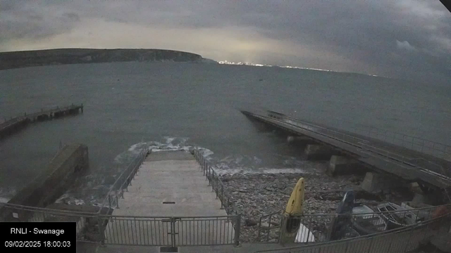 A coastal scene at dusk, showing a rocky shore with a concrete ramp leading down to the water. On the left, a curved pier extends into the choppy sea, while a small section of a boat ramp is visible. In the foreground, there are some scattered rocks and a yellow kayak resting on the beach. The sky is overcast, with dark clouds and a faint glow of lights on the distant horizon, possibly from a town or city.