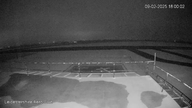 The image shows a dimly lit outdoor scene from a webcam positioned at the Leicestershire Aero Club. A large, illuminated area is visible in the foreground, possibly a runway or helipad marked with an "H" shape. There is a railing visible along the edge of the platform, and the dark sky suggests it is either dusk or nighttime. In the distance, faint lights can be seen on the horizon, indicating the presence of buildings or vehicles. The overall atmosphere is quiet and serene, with a sense of stillness in the air. A timestamp in the top right corner indicates the date and time as February 9, 2025, at 6:00 PM.