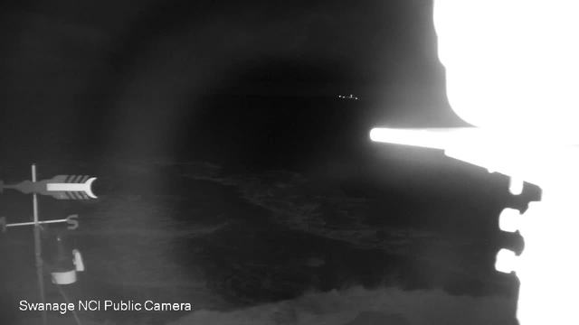 A black and white image showing a coastal scene at night. The foreground features a weather vane with directional indicators, while the background displays dark water with faint glimmers of distant lights on the horizon, suggesting boats. The overall atmosphere is dark with low visibility.