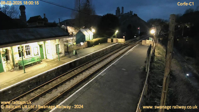 A dimly lit railway station platform is shown with stone walls and a green bench next to a row of windows. On the left, a small building with a green roof is partially obscured by trees. There is a signpost reading "WAY OUT" visible on the platform. The railway tracks are in the foreground, leading into the darkness. In the background, a silhouette of a castle or ruins can be seen on a hill, under a cloudy sky. The overall scene conveys a tranquil yet slightly eerie atmosphere of twilight.