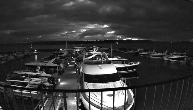 A nighttime view of a marina filled with numerous boats docked side by side. The scene is illuminated by some ambient lights on the boats and mooring posts, revealing their shapes and outlines against a dark, cloudy sky. The water appears calm, reflecting faint light and the silhouette of the shoreline in the background. The image is primarily in black and white, enhancing the contrast between the boats and the dark surroundings.