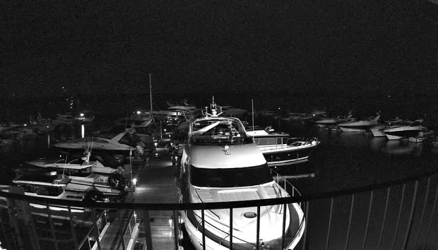 A nighttime view of a marina filled with various boats and yachts. The scene is illuminated by soft lights reflecting off the water. In the foreground, a large yacht is moored at a dock, and several smaller boats are visible nearby. The overall atmosphere is calm and tranquil, with the darkness of night surrounding the area.