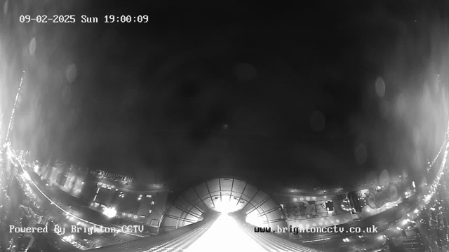 A black and white webcam image showing a view from a high point, looking down at a city scene below. The lower part of the image features illuminated streets with lights suggesting buildings and vehicles. The upper portion is mostly dark with some light spots, indicating potential clouds or atmospheric disturbances. A time and date stamp is located at the top, indicating Sunday, February 9, 2025, at 19:00:09. The bottom left corner has text stating "Powered By Brighton CCTV" and the website address "www.brightoncctv.co.uk."