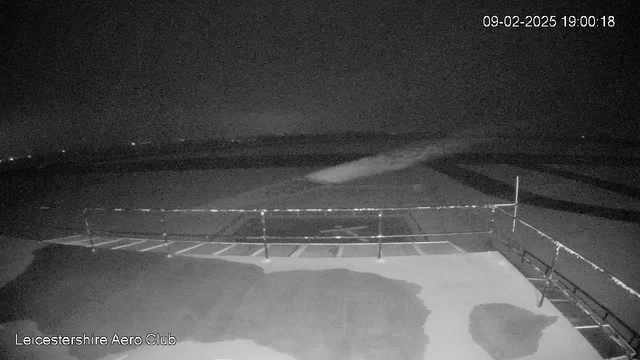 A dark scene is captured from a webcam at the Leicestershire Aero Club. In the foreground, there's a flat surface that appears wet or reflective, possibly a runway or patio area. A railing runs along the edge of the surface. In the background, the sky is mostly black with some faint lights indicating distant sources, but no clear outlines or features are visible. The image is low in contrast and lacks distinct details due to the lighting conditions. The timestamp indicates the date and time of the capture.