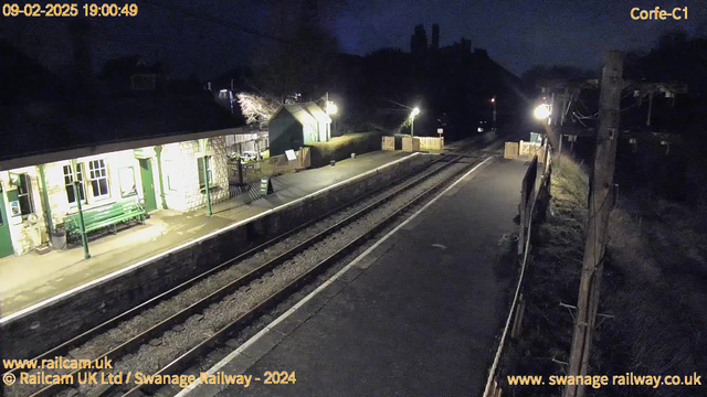 A dimly lit railway station at night. The stone building on the left features green accents and a bench in front. The platform is empty, with two railway tracks visible leading into the distance. There are wooden barriers at both ends of the platform, and a small sign indicating the exit. To the right, a faintly lit pole is visible, surrounded by dark areas and sparse vegetation. The sky is overcast, contributing to the somber nighttime atmosphere.