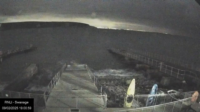 A dark, cloudy scene depicting a coastline at night. In the foreground, a series of steps lead down to a rocky area by the water, enclosed by a metal railing. Two kayaks, one yellow and one blue, are positioned nearby on the ground. In the distance, a faint outline of a hill can be seen against the sky, which has a slight glow from lights beyond the horizon. The overall atmosphere is calm with no visible movement on the water.