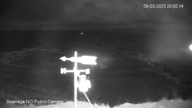 A dark, nighttime seaside view captured by a webcam. In the foreground, a weather vane with directional arrows indicating North and South is visible. The background features the dark surface of the sea, with faint waves and a few distant lights on the horizon. The upper right corner displays the date and time: September 2, 2025, at 20:00:14.