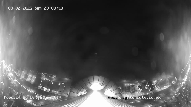 A black and white webcam image showing a cityscape at night. The view is directed down a street lined with bright lights, indicating activity. The sky appears dark with a slight haze. There is a prominent structure in the center that is illuminated, possibly a roof or archway, leading the eye along the street. The date and time are displayed at the top, along with a watermark at the bottom for the CCTV service.