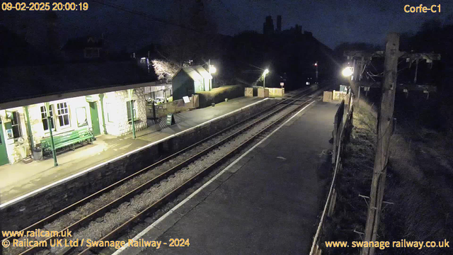 A dimly lit train station at night, featuring a stone-built platform on the left with a green wooden bench and a small green building. The platform is lined with railway tracks that lead into darkness. There is a wooden fence and a sign that reads "WAY OUT" near a small outdoor area. A light pole casts a soft glow along the platform, while the background features trees and possible buildings, partly obscured by darkness.