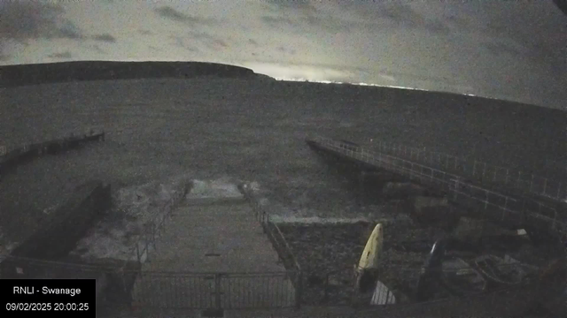 A dark, cloudy scene captured from a webcam, showing a waterfront area during the evening. The foreground features a partially visible pier extending into the water, with some waves lapping at the edge. On the right, there is a yellow object, possibly a kayak, resting on the ground. The background includes a distant coastline, barely illuminated against the dark sky. The overall atmosphere is tranquil yet somewhat ominous due to the low light conditions.