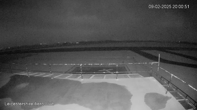 A dark, nighttime view of a runway at Leicestershire Aero Club. The image is mostly in shades of black and gray, with a small section of white light reflecting on a wet surface. A railing is visible in the foreground, and the runway has markings that resemble a helicopter landing pad. In the distance, faint lights can be seen, likely from nearby structures or vehicles. The time is recorded as 20:00:51 on February 9, 2025.